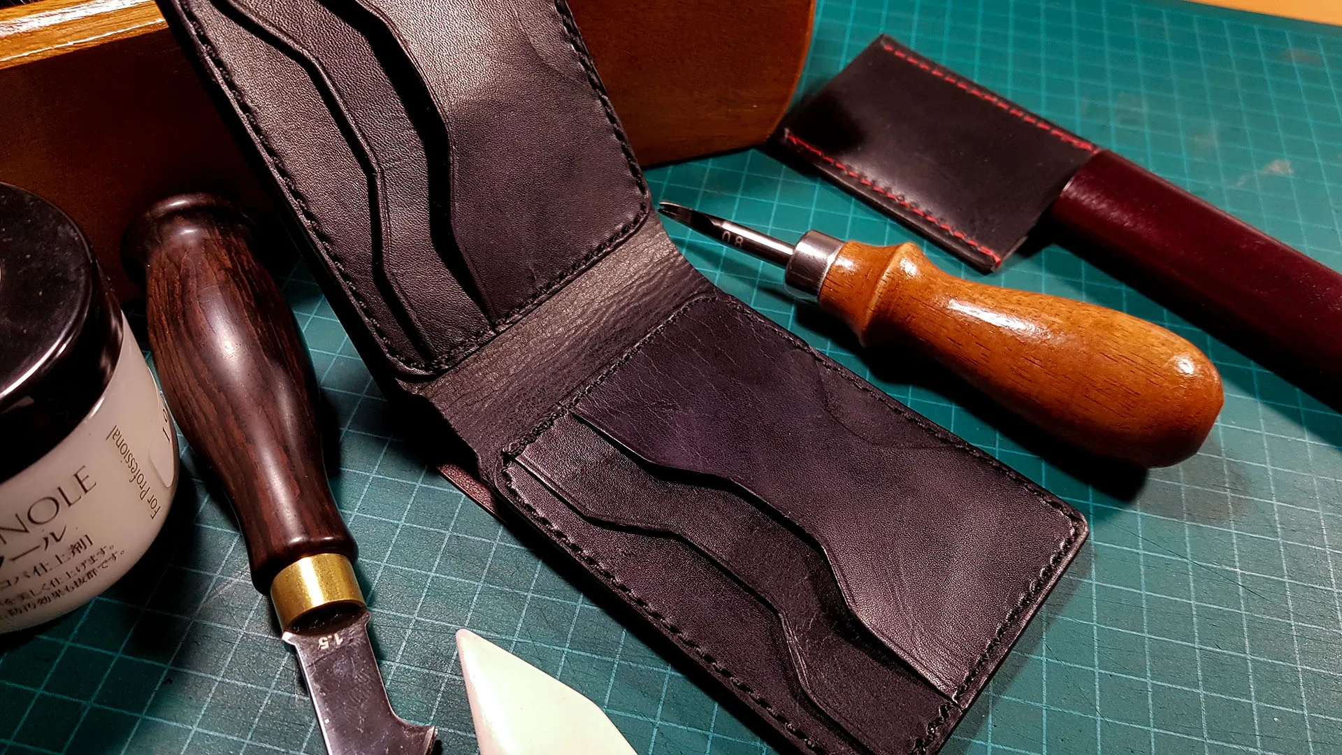 Open black bifold wallet on a cutting mat, surrounded by leather-working tools