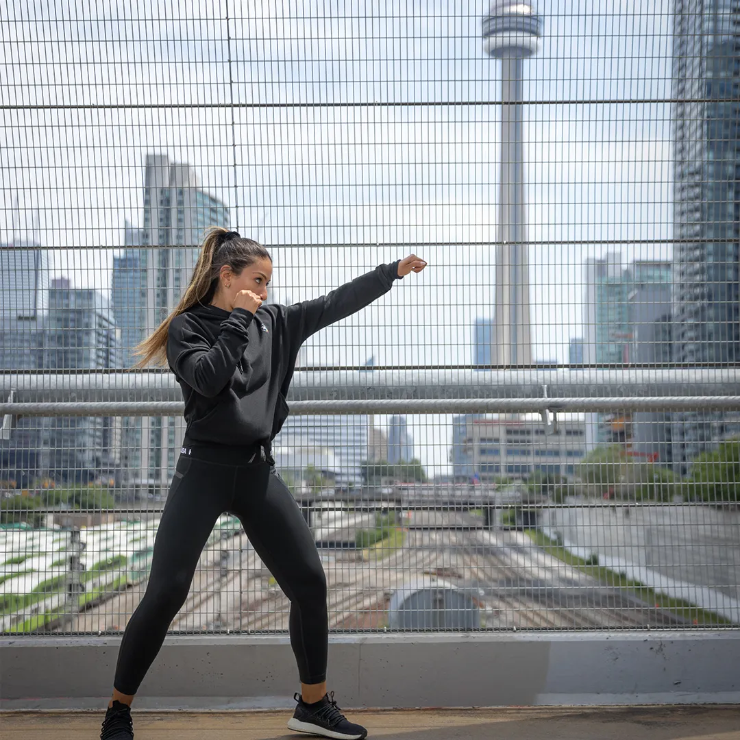 Woman in a black Hayabusa jacket throwing a high kick on a rooftop with the skyline behind