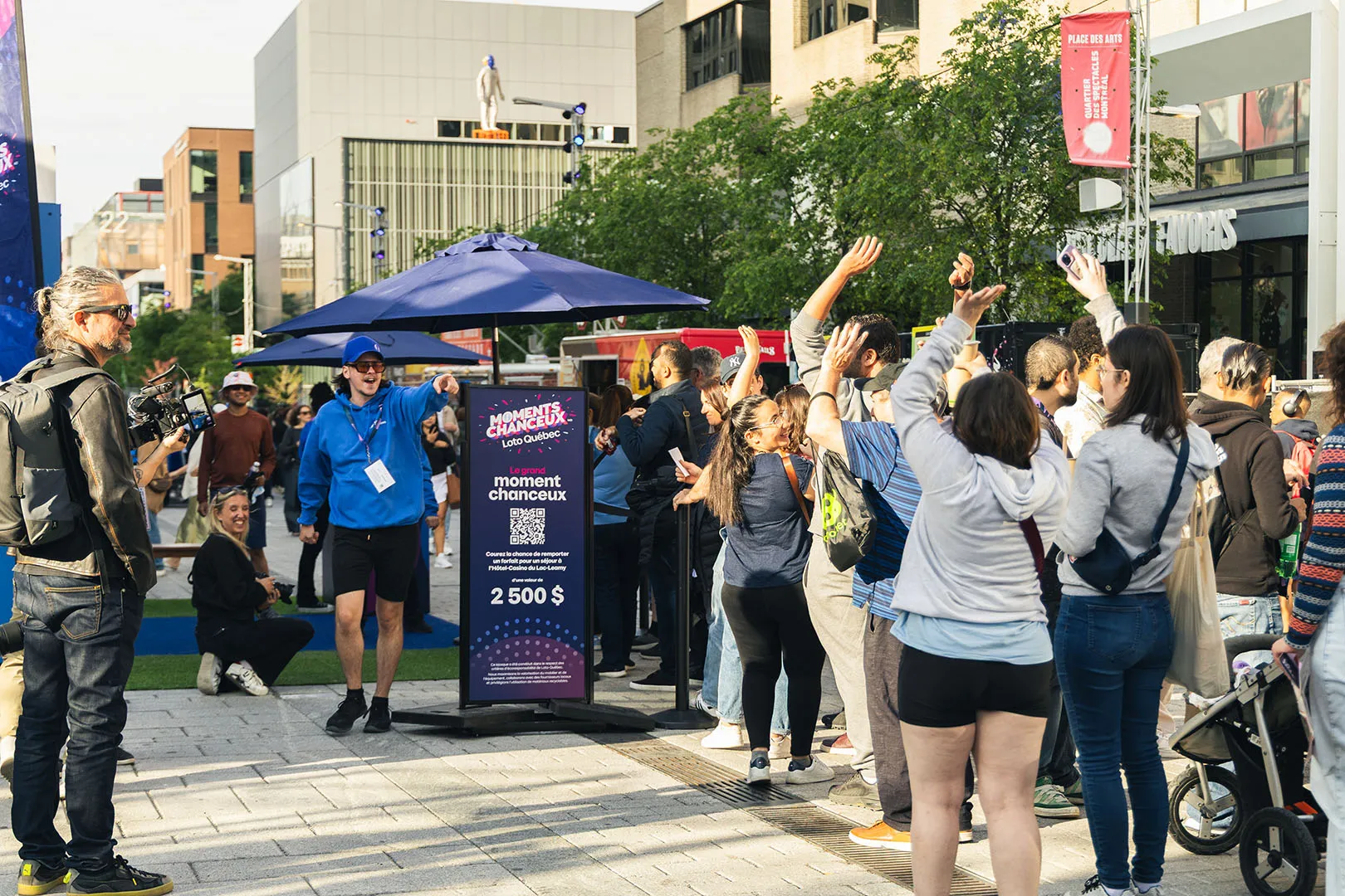 Festival crowd with raised hands around a blue kiosk displaying Le Grand Moment Chanceux with $2500 prize signage