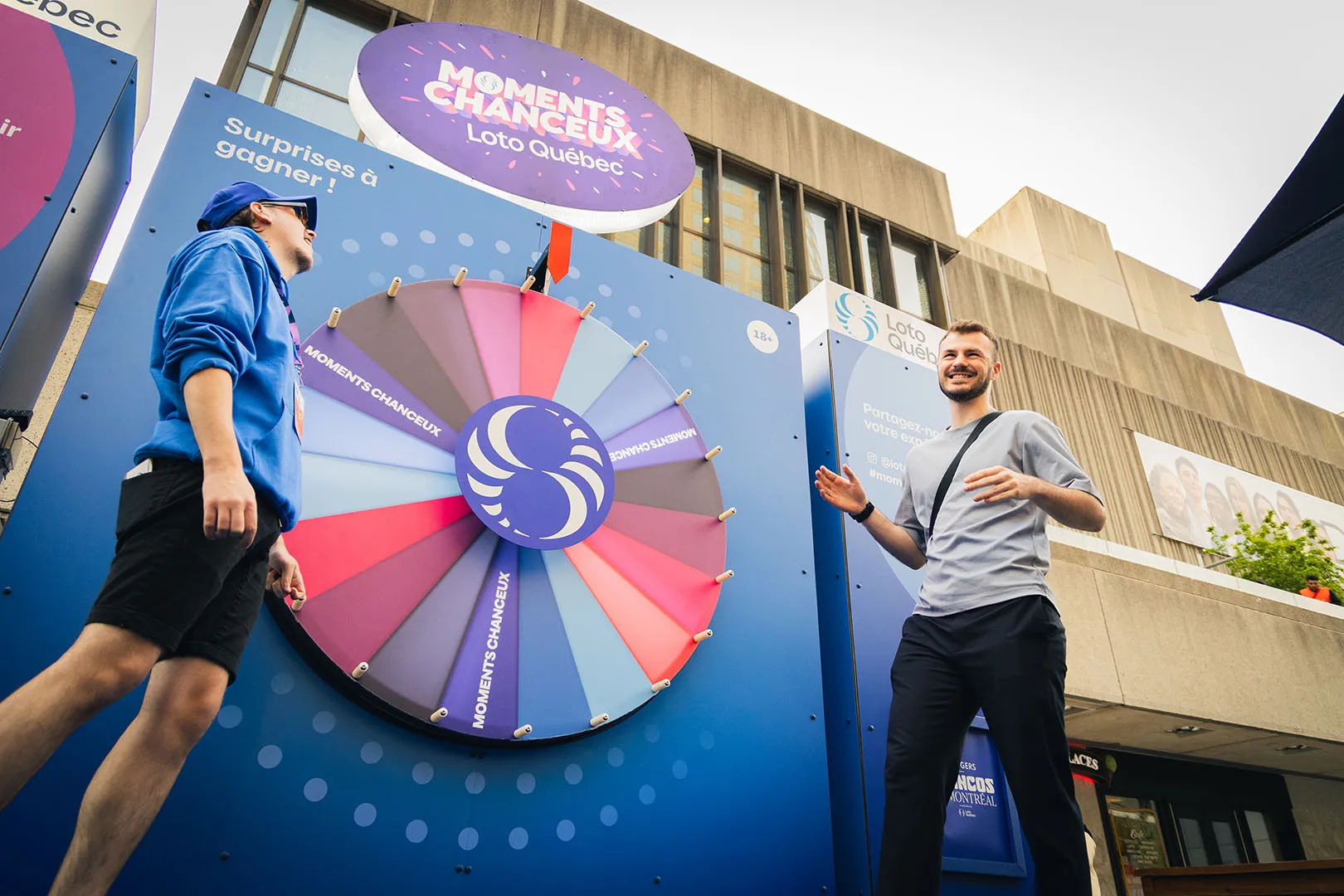 Two participants interacting with the multicoloured Roue des Moments Chanceux wheel on a blue branded backdrop
