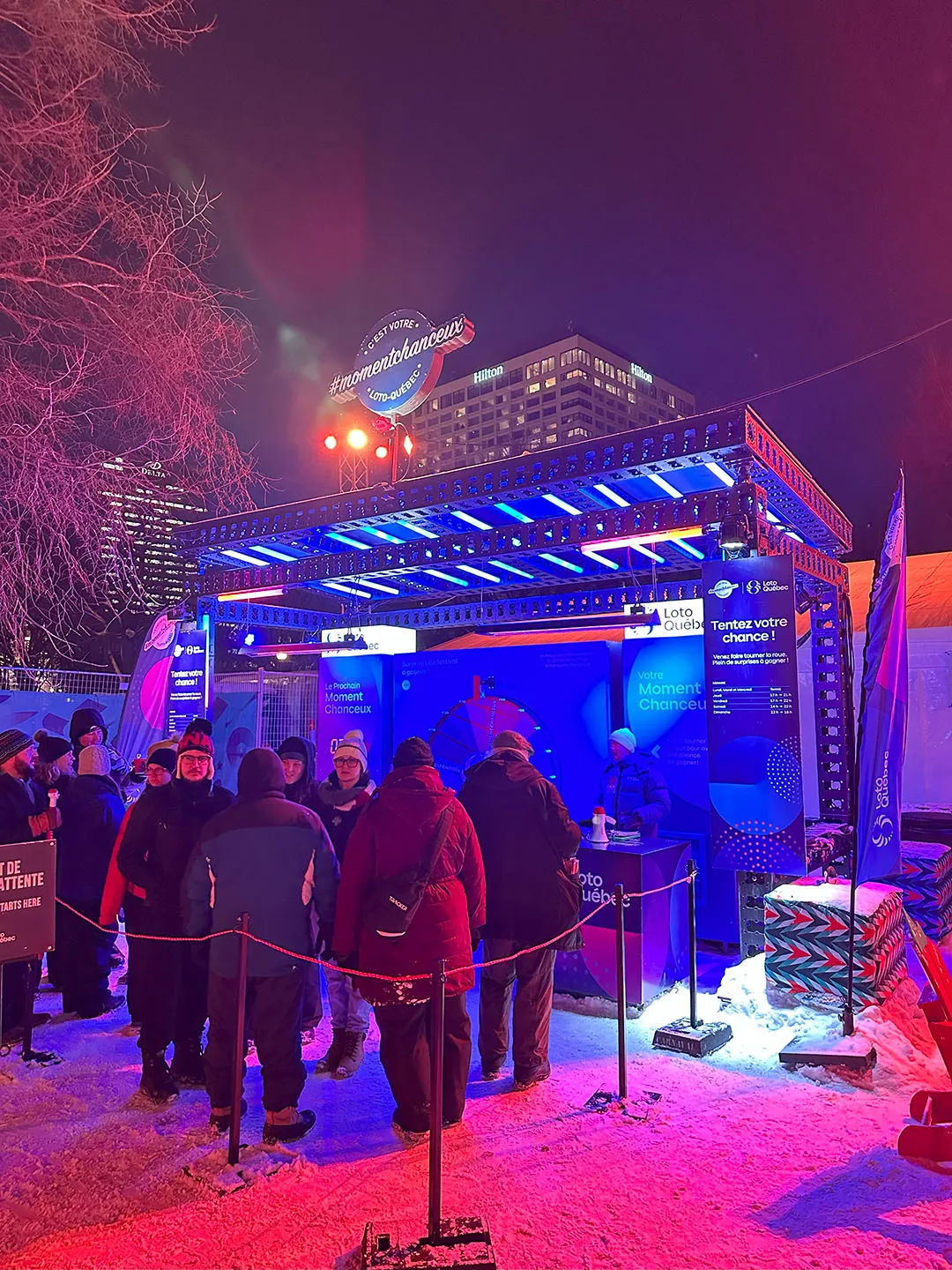 Winter festival venue at night with blue LED beams overhead, red neon accents, and visitors in winter clothing