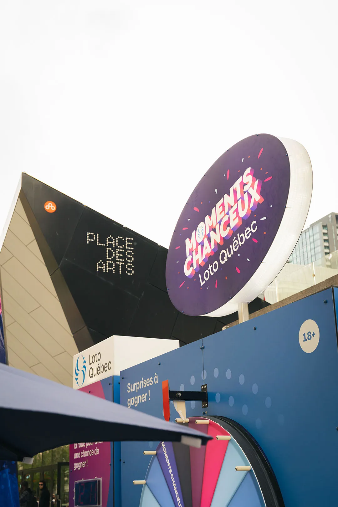 Exterior view of the Roue des Moments Chanceux wheel signage at daytime with Place des Arts in background
