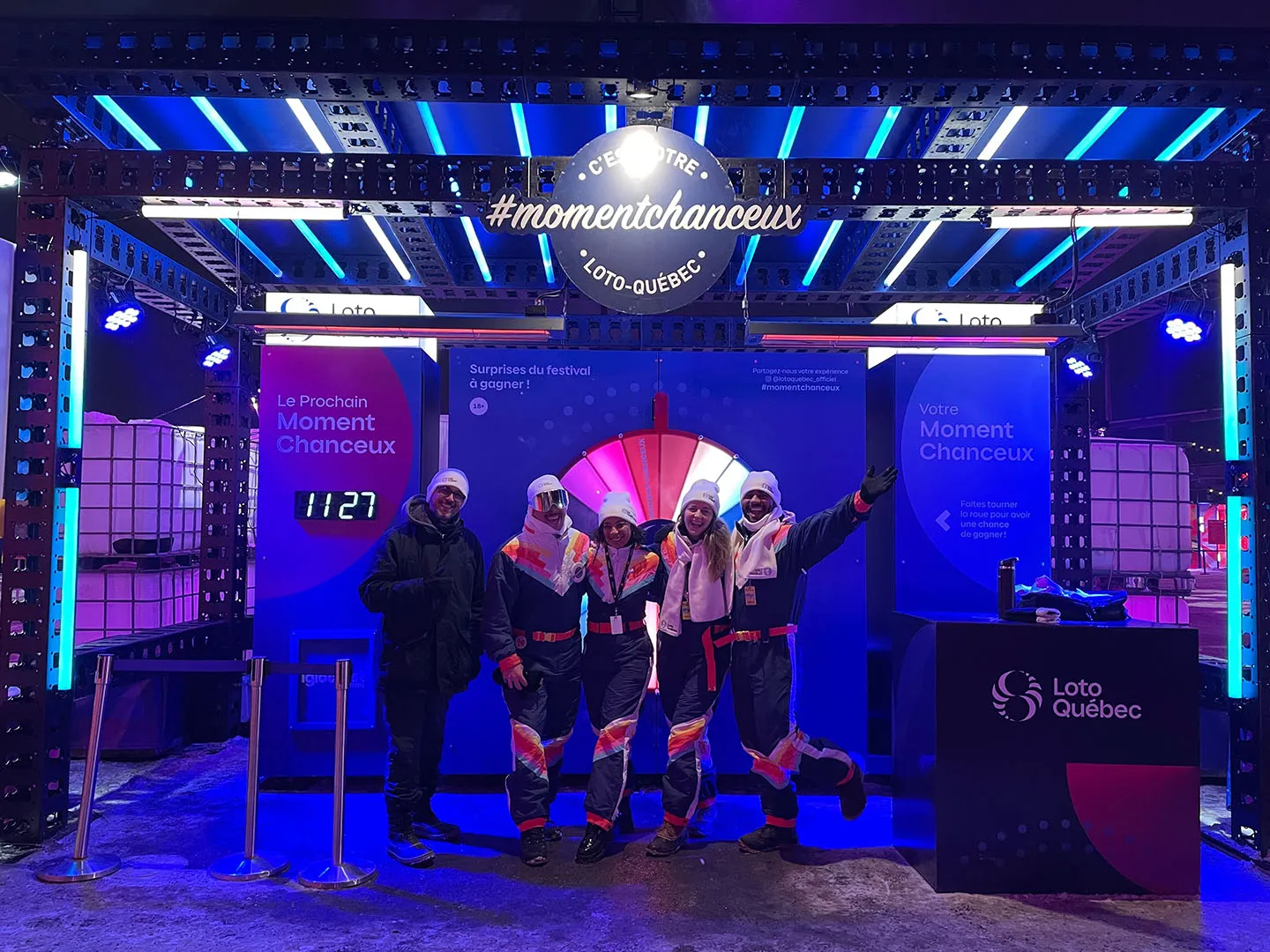 Team posing before the Roue des Moments Chanceux wheel indoors under blue LED grid ceiling with countdown timer