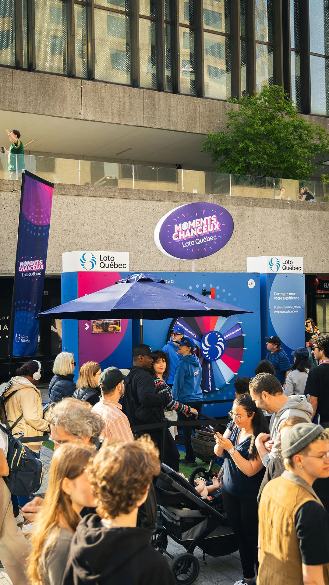 Daytime urban plaza crowd gathered around the Roue des Moments Chanceux booth with wheel and signage overhead