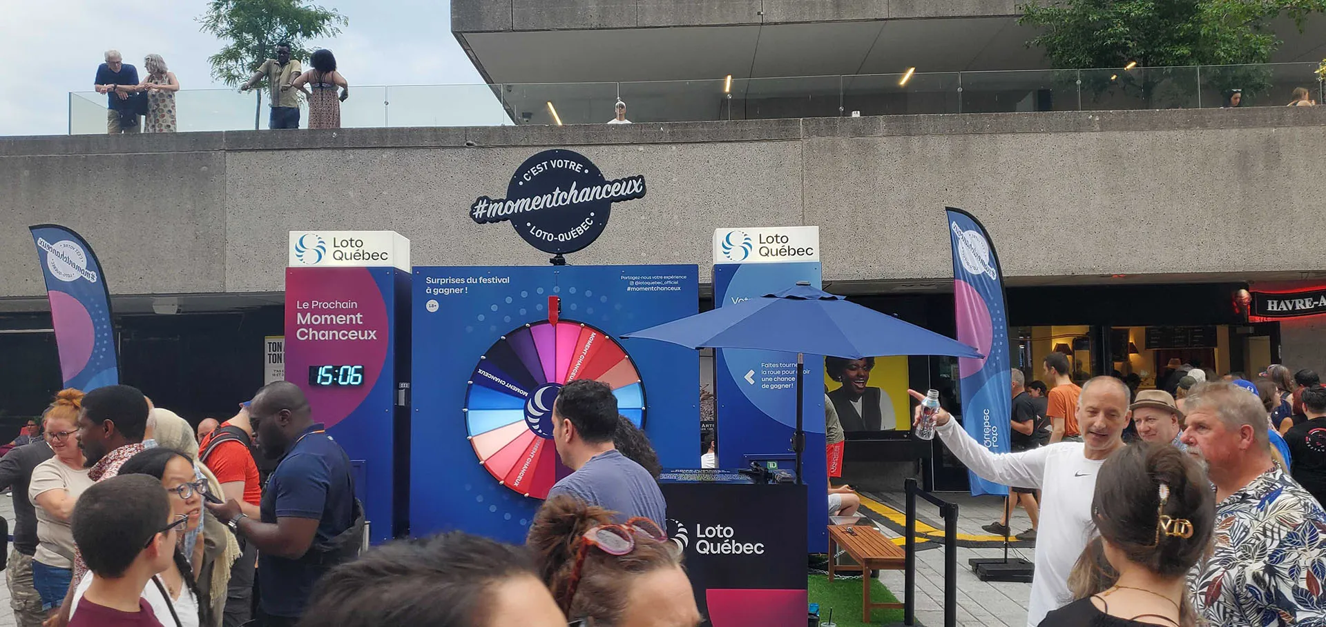 Daytime festival plaza with large crowd around wheel setup, countdown timer, and promotional banners visible