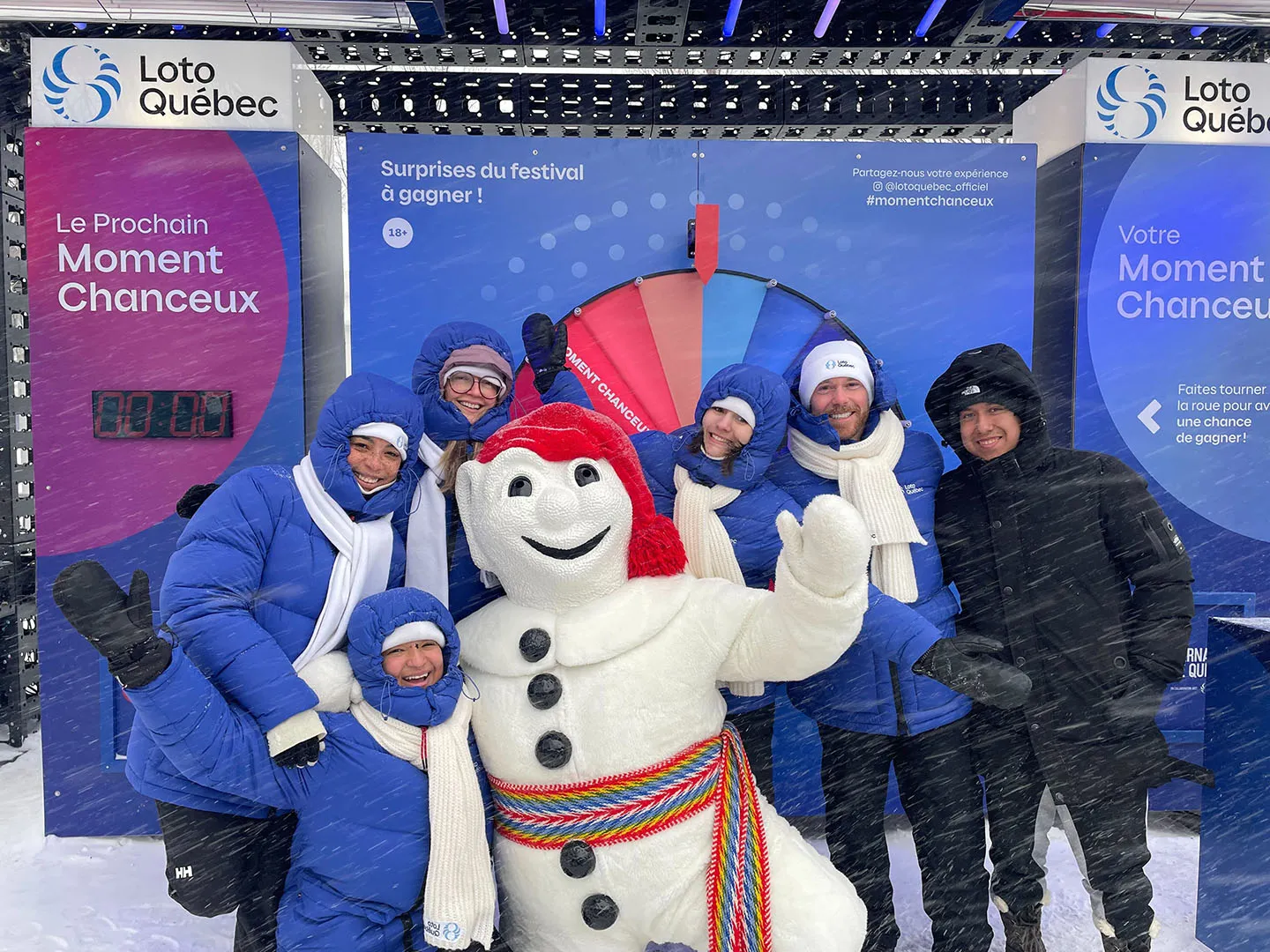 Snowman mascot in red hat posing with team staff in blue winter outfits at festival activation