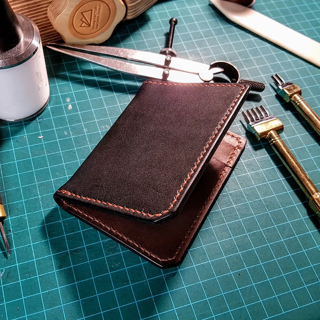 Hand-stitched dark brown card wallet on a cutting mat, surrounded by leather tools