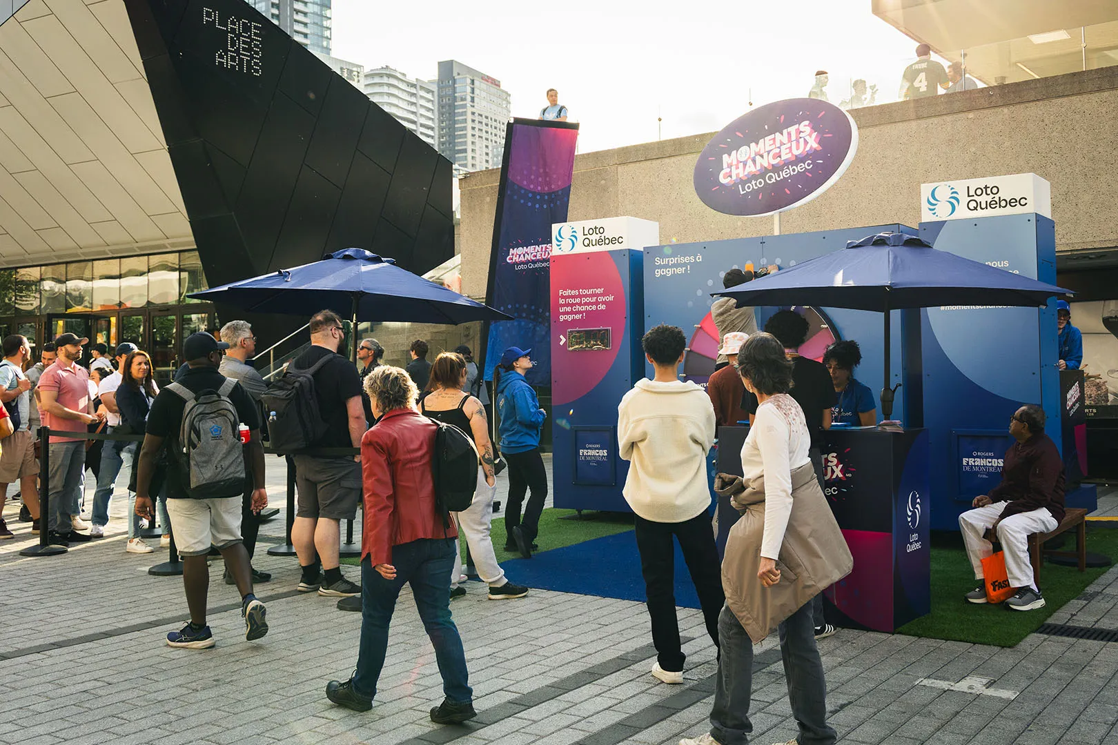 Daytime festival plaza with blue umbrellas, overhead pink signage, and a branded kiosk